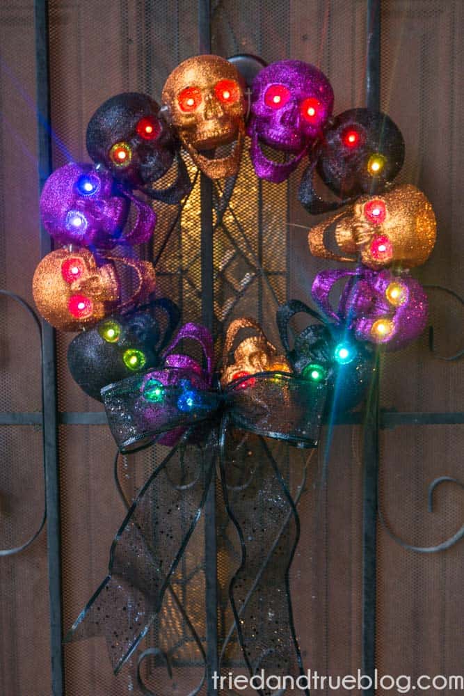 Metal door with a dollar store wreath made of glitter purple, orange, and black skulls. Eyes have different colors of lights shining.