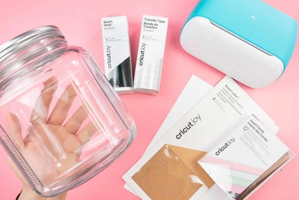 Hand holding a glass jar with Cricut Joy and supplies in background.