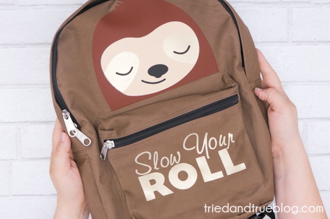 Hands holding brown backpack with vinyl sloth and the words 