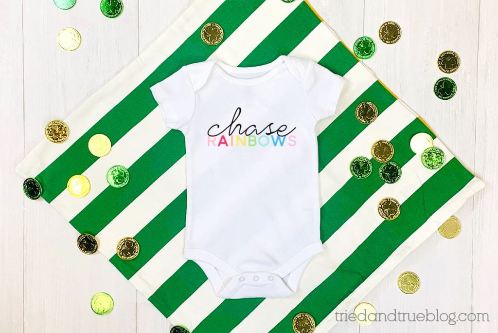 White baby onesie on green/white striped background with the words "Chase Rainbows."
