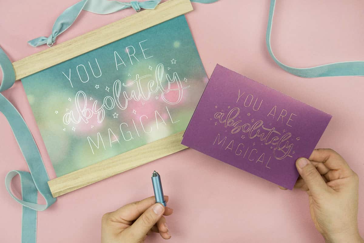 Hand holding Cricut Foil Transfer Tool with two varieties of the You Are Magical Single Line Free SVG.