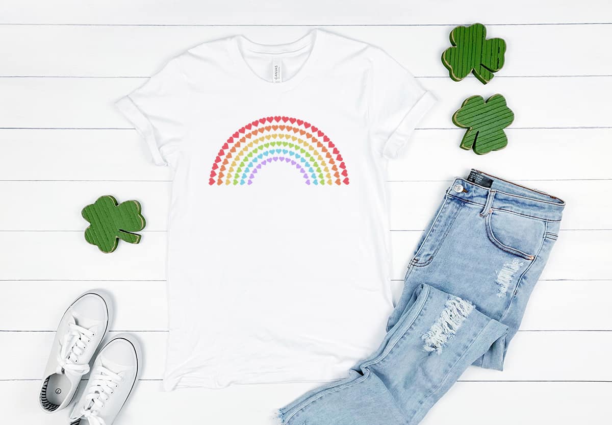 White t-shirt on shiplap background with jeans and shamrocks around it. T-shirt has St. Patricks Free SVG Heart Rainbow on it.
