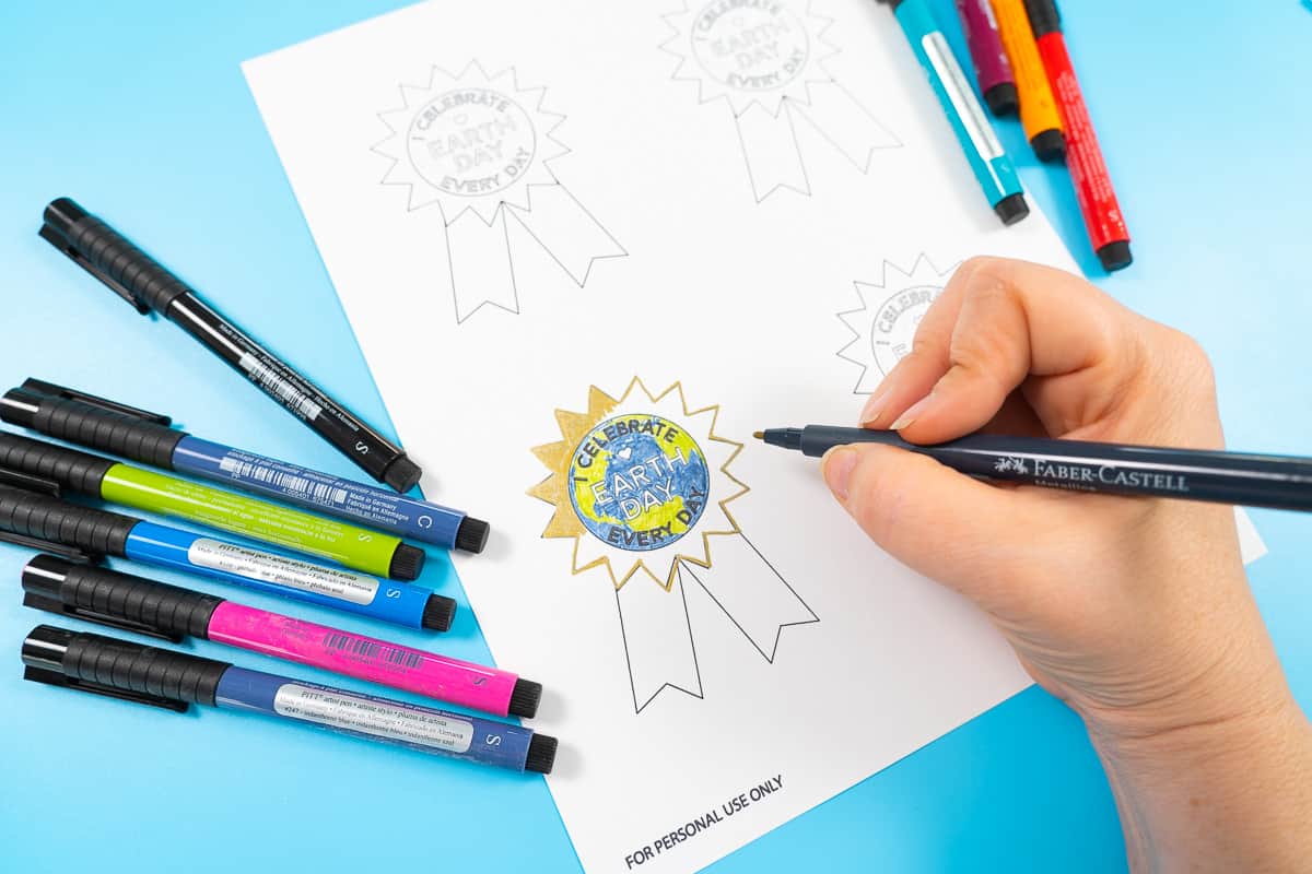 Hand holding marker on blue background while coloring in Earth Day free printable badges.