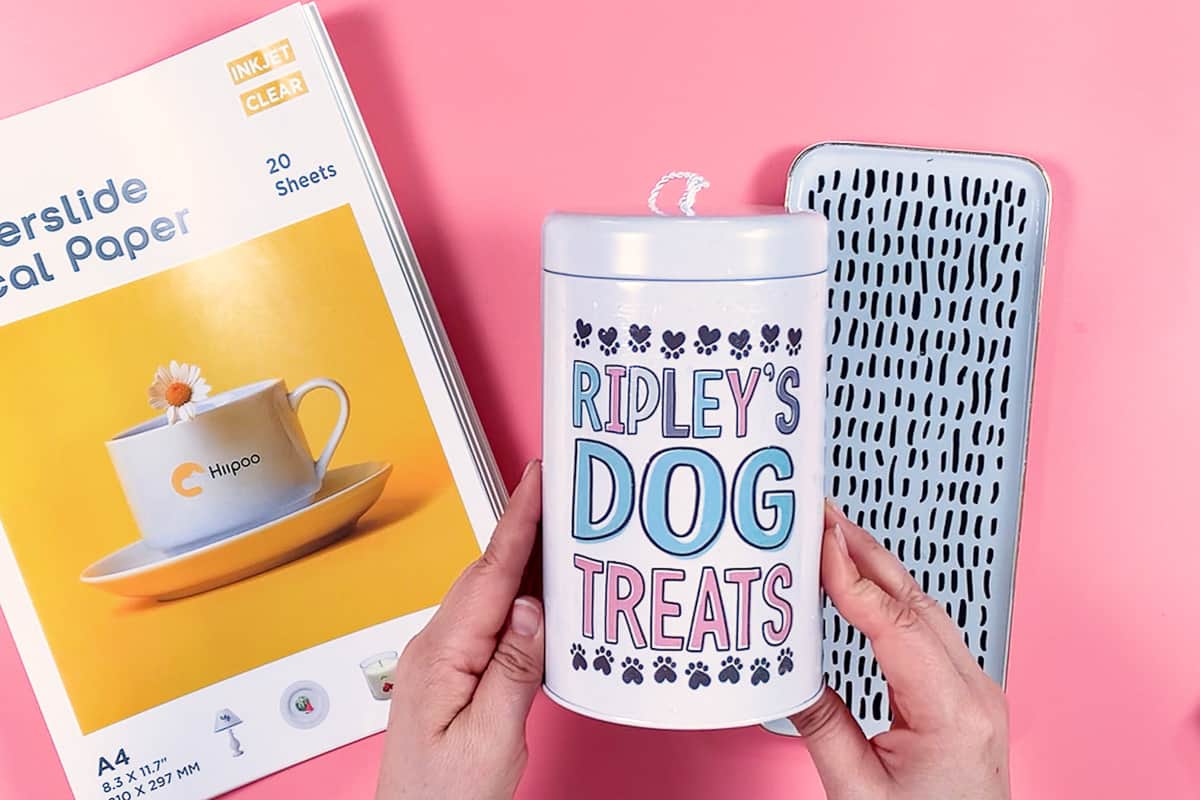 Hand holding a dog treat holder with words "Ripley's Dog Treats" on a pink background with waterslide decals paper.