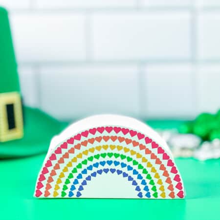 a paper rainbow St. Patrick's Day Gift Box on a green table next to a st patrick's day hat.