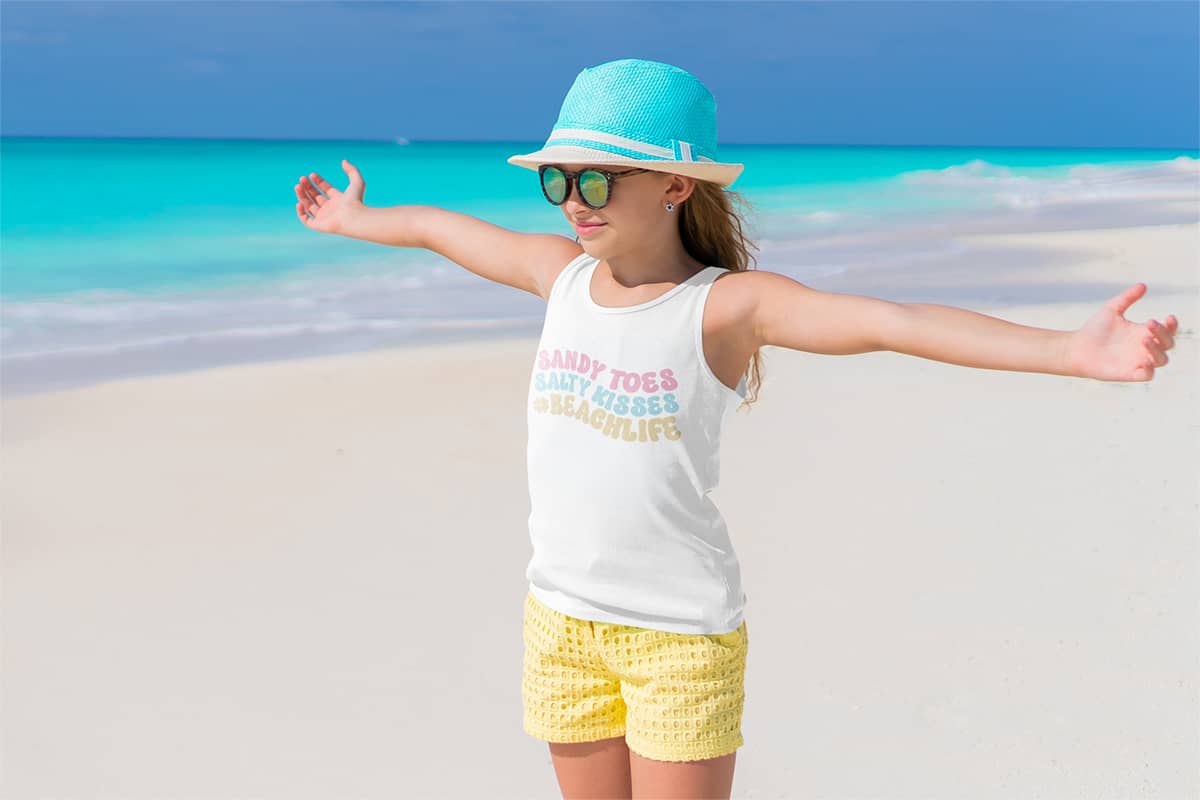 A young girl standing on the beach with her arms outstretched wearing a tshirt with Sandy Toes Salty Kisses Free SVG File