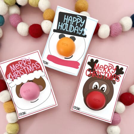 Three Christmas Lip Balm Free Printables on a pink background with pom poms.