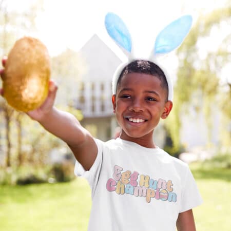 A boy wearing bunny ears holding a golden egg with white tshirt that has the Egg Hunt Champion free svg file on it.