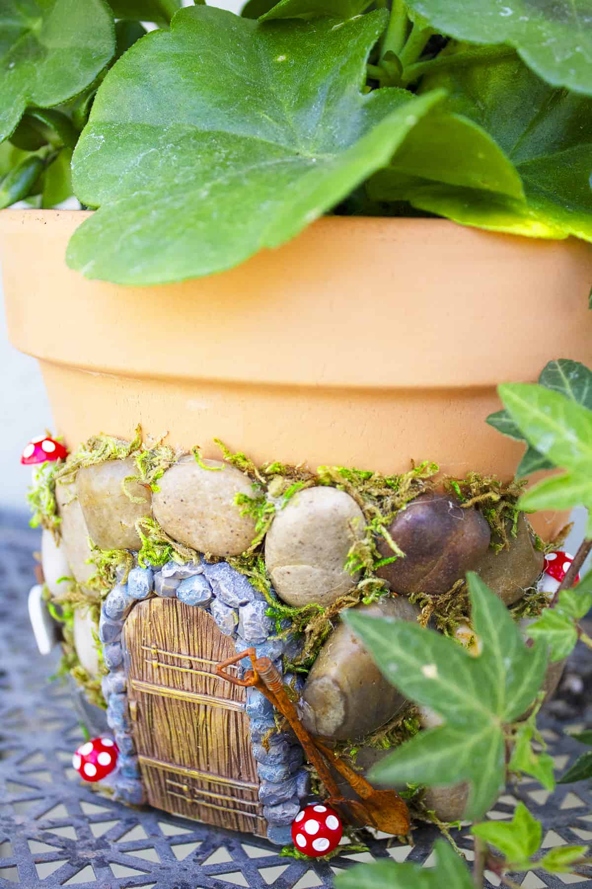 Terracotta flower pot decorated with a fairy garden theme, featuring a small door and mushrooms, nestled among green leaves.