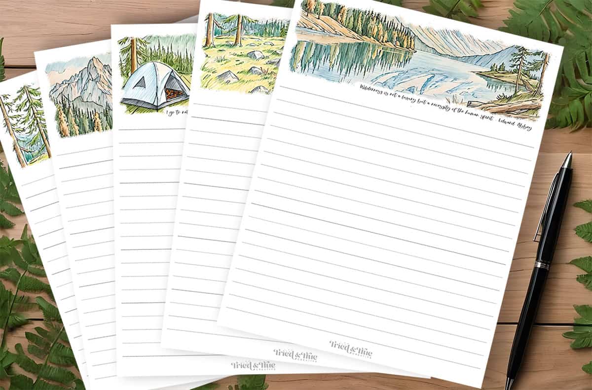Four lined Camping Journal Pages featuring different outdoor-themed illustrations at the top, including a tent, mountains, and a lake. A pen and fern leaves are placed around the notepads.
