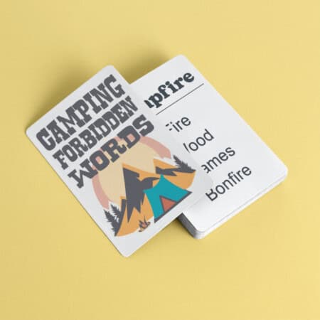 A set of cards on a yellow surface, with one card displaying "CAMPING FORBIDDEN WORDS" and an illustration of a tent and campfire, and another card listing words like "fire," "wood," and "flames.