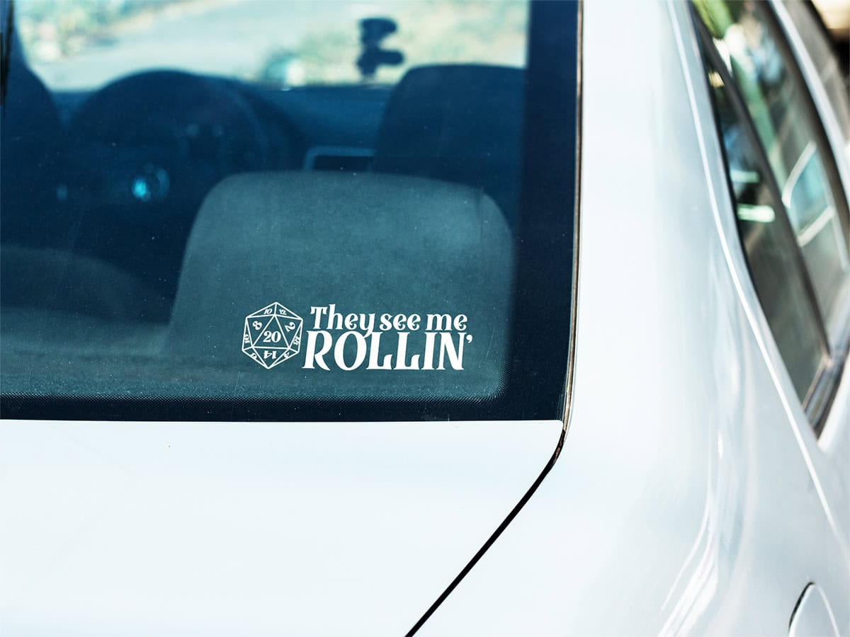 A white car's rear window displays a sticker with a 20-sided die and the text "They see me ROLLIN'.