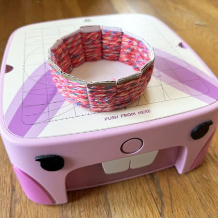 A colorful, woven bracelet made using cardboard weaving sits on top of a pink crafting machine with a cartoon face design, placed on a wooden floor.